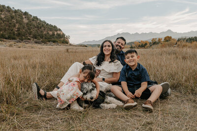 A family of four and their dog sit smiling together in a grassy field with mountains in the background—perfect playful family photos in Montrose, Colorado.
