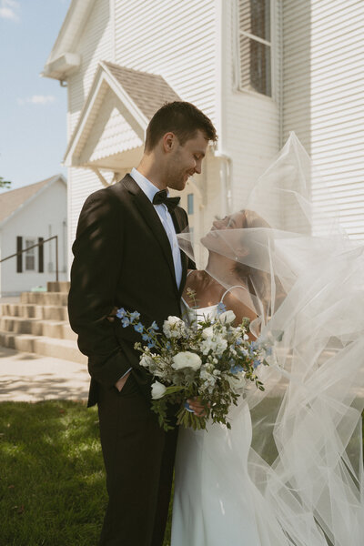 Man in tuxedo looking down and smiling at women in wedding dress smiling up at man while holding bouquet of flowers and her vail is covering her face while flowing in the wind