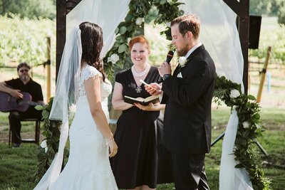 Iranian Bride in a long white dress and veil and Irish Groom in a black suit and tie stand before the officiant dressed in black under the arbor decorated with white drapery, green garlands and white flowers