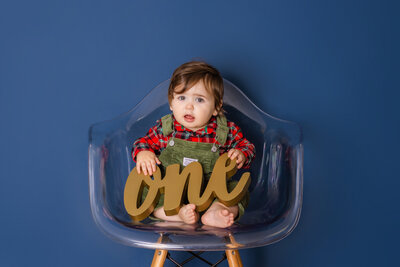 Cake Smash Photographer, a baby girl sits on a flowery chair and holds a wooden carving that reads "one"