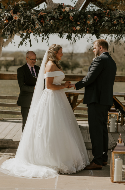 Bride and Groom exchanging vows under outdoor wedding pergola at Eden Barn Cumbria Lake District with flower arch flower installation apricot flowers pampas eucalyptus