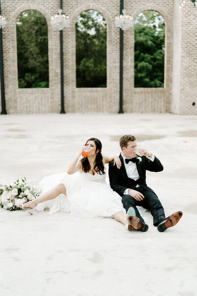 bride & groom celebrate getting married by chugging a drink while sitting on the ground
