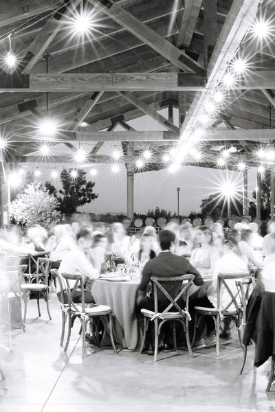 black and white image of wedding reception group seated at tables in large, barn-like venue.