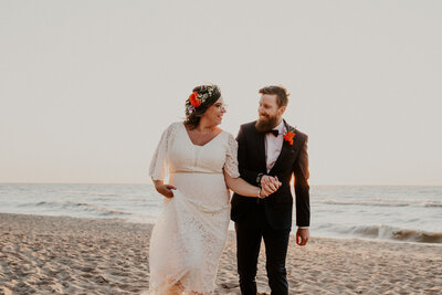 Bride and Groom walking on Grand Bend beach at sunset for portraits. Bride and groom holding hands, smiling and looking at each other. Bride is holding her dress up with the other hand.