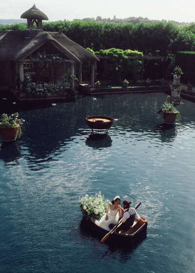 Bride and groom rowing across a romantic garden pond in a small boat, captured in a dreamy, cinematic style by a luxury documentary wedding photographer UK.