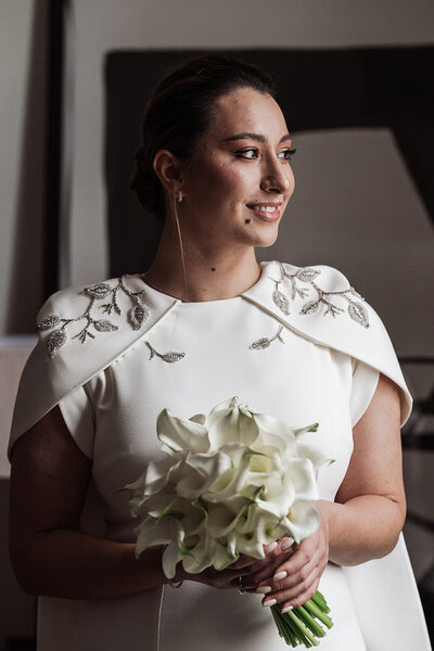 Smiling bride wearing wedding dress with embroidered shoulders and cape attachment holding a bouquet of white calla lilies