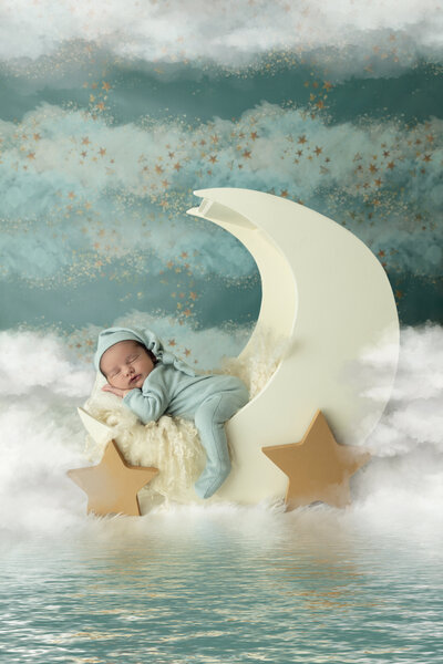 A newborn baby sleeps in a matching onesie and night cap on a moon shaped bed floating in the clouds above the ocean posed by a New Jersey Newborn Photographer