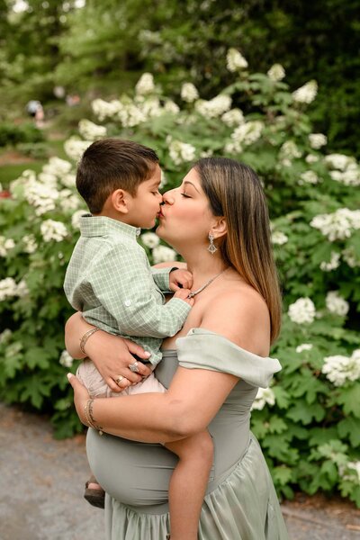 mom to be holds her toddler son giving him a kiss  as he sits on her bump
