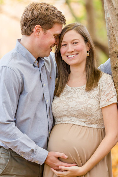 Husband embraces his smiling wife as she embraces her pregnant belly during maternity photo shoot