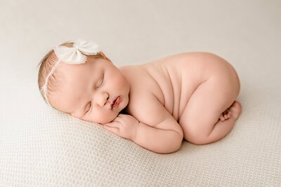 Newborn baby girl is posed on a cream colored background wearing a cream color bow with her hand underneath her cheek naked and curled up bows gently during her Studio newborn session