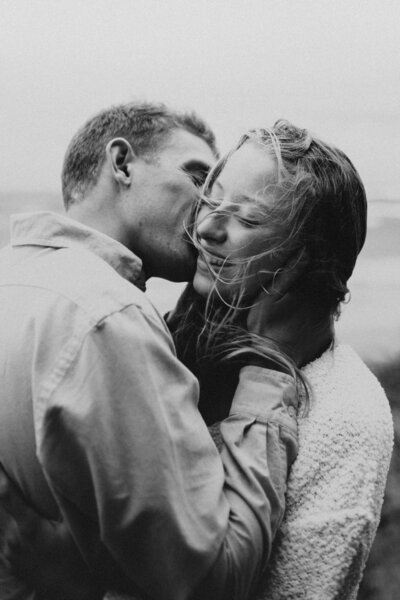 A man kissing his fiancé on the cheek on a windy day while her hair blows in her face
