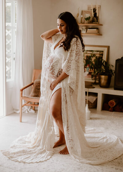 Pregnant woman in white lace dress posing for maternity photoshoot in her home