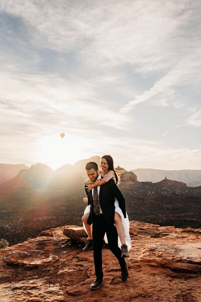 groom giving bride piggy back ride at sunrise in sedona az while hot air balloons rose in the background