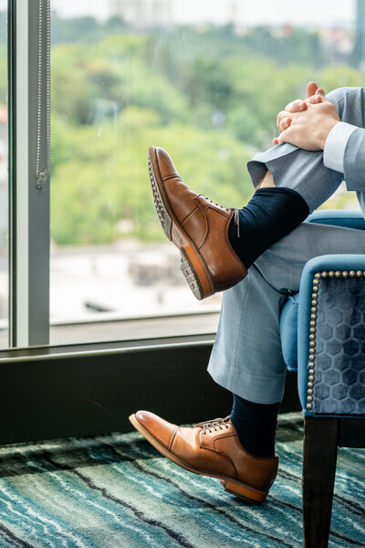 Groom wearing brown leather shoes relaxing by large window – photographed by wedding photographer Niko Coric – Lumen Clarity Media