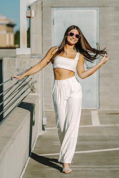 Brunette senior in white crop top and white pants, standing in parking garage