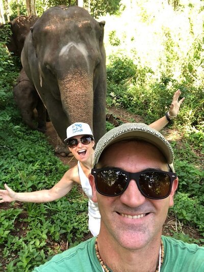EnForma co-founders Alicia and Errin smiling beside an elephant in a lush Thai forest, capturing a moment from their wellness journey in Thailand that reflects global exploration, mindfulness, and connection to nature.
