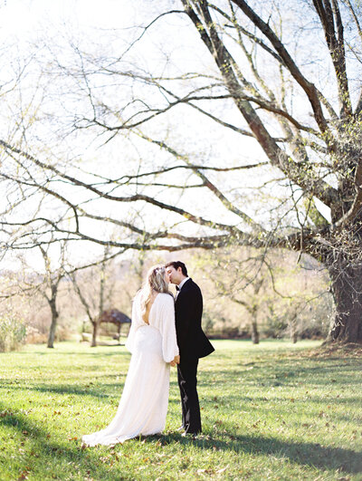 A just married couple shares a kiss under some large trees. The bride is wearing a long sleeved white gown and the groom wears a tuxedo.