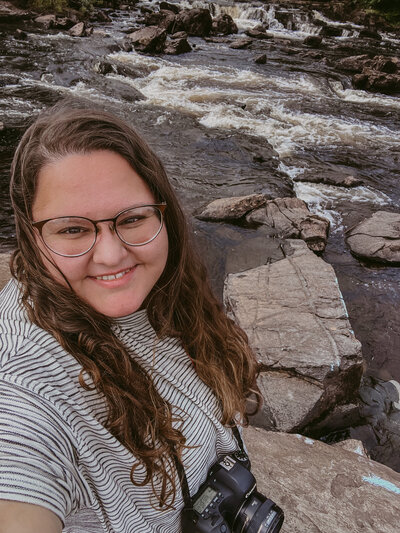 Plus size brunette woman with glasses and striped shirt and dslr canon camera takes a selfie while standing in the falls of dochart in scotland