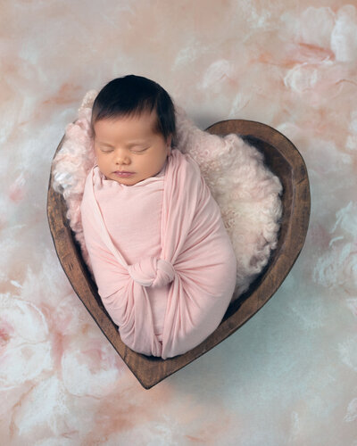 newborn girl wrapped in pink sleeping in a heart shaped bowl