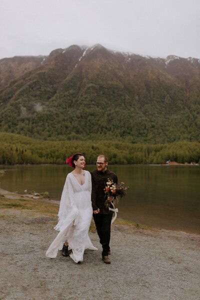 A bride and groom walk and laugh together next to Mirror Lake in Alaska during their Alaska elopement. A tall snow-capped mountain is in the backgorund.