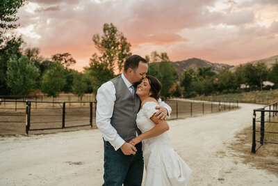 A newlywed couple shares a tender moment outdoors at dusk, with the groom dressed in a gray vest and white shirt, embracing the bride who is radiant in an off-the-shoulder lace wedding gown. They are smiling joyfully, with the groom leaning in for a kiss on the bride's forehead. The backdrop features a dramatic sunset sky in warm hues, with silhouettes of mountains and trees, adding to the romantic ambiance of the scene.