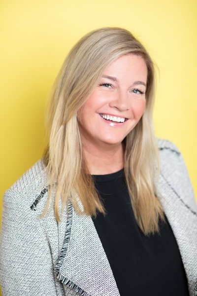 Woman smiling wearing a gray jacket and blonde hair with a yellow background