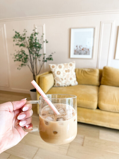Hand holding a clear mug of iced coffee with a straw with a couch in the background