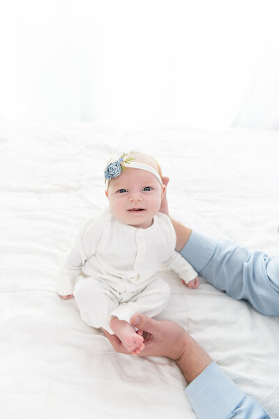 A newborn baby girl is held up by dad on a bed in a white onesie and blue flower bow posed by an Atlanta newborn photographer