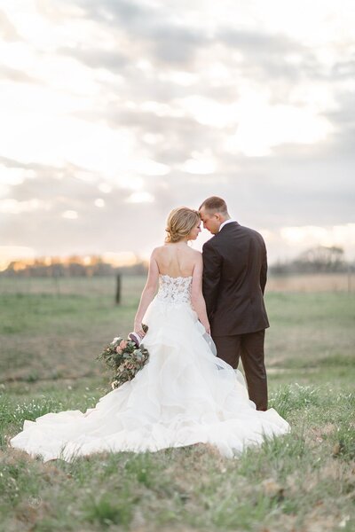 A bride and groom standing in a field at sunset.  They are facing each other and the bride has her bouquet at her side