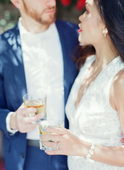 Man in blue suit and white shirt and lady in white gown and red lipstick come together for a drink of whisky.