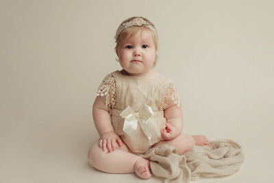 sad looking baby on a white drop wearing a white lace romper