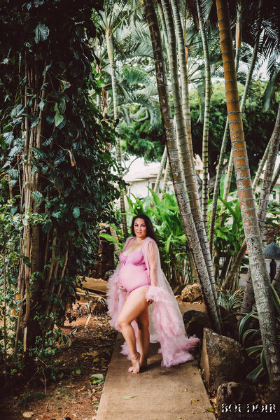 A glamorous pregnant woman stands in lush  tropical setting wearing lingerie and pink robe