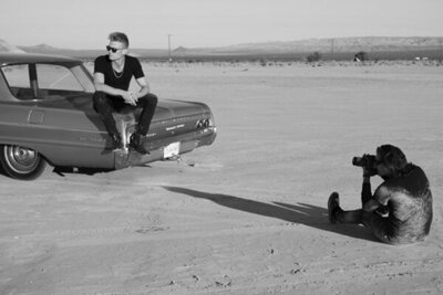 Behind the scenes photo Mark Maryanovich on ground in desert photographing musician Ryan Guldemon as he sits on trunk of vintage car wearing sunglasses and looking over his shoulder black and white image