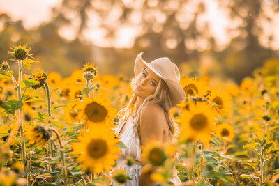 blonde senior girl with a cowboy hat on in a field of big yellow sunflowers at sunset