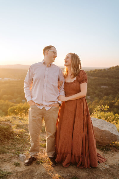 A couple stands on a mountaintop at sunset, smiling at each other. The woman wears a rust-colored dress and holds the man’s arm as golden light bathes the scenic landscape behind them.