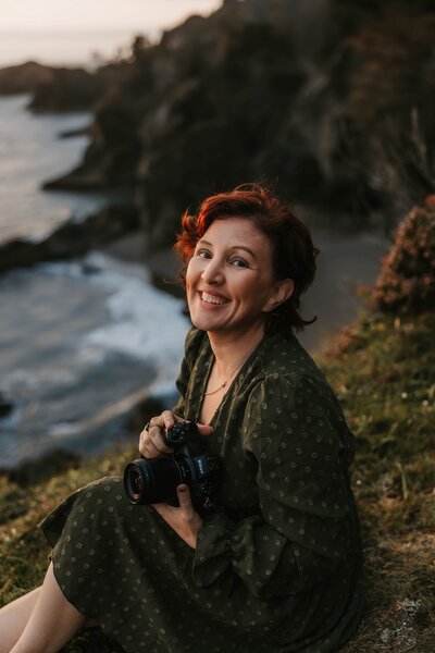 Woman with short red hair sits on a grassy cliff by the ocean, smiling and holding a camera—capturing moments at an Oregon coast photography retreat for wild-hearted artists.
