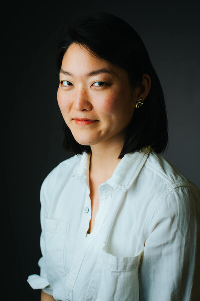 Studio portrait with grey backdrop of a young Chinese-American woman in a casual white-button-up shirt and spiky brass earrings. Her black hair is shoulder-length and one side is tucked behind her ear.