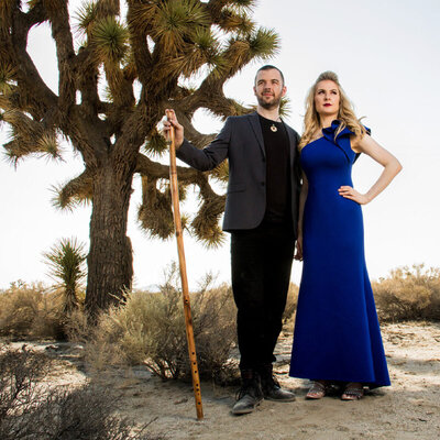 Musician Portrait Duo standing next to joshua tree in desert Jason Campbell holding long wood flute hand Robin Campbell in blue dress with on hip