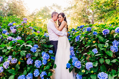 A bride and groom embracing each other standing among a bunch of plants with blue flowers