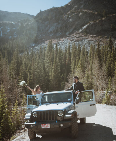 Couple standing out opposite doors of their jeep cheering and smiling at each other. the bride is holding her bouquet, and the groom is wearing a black suit. the jeep is silver. they are in the breckenridge mountains after their hiking elopement.