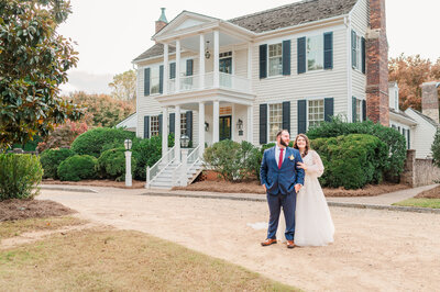 A brunette bride holding the arm of her new husband in a blue suit in front of The Sutherland in Raleigh on a fall afternoon enjoying their Raleigh wedding photography by JoLynn Photography