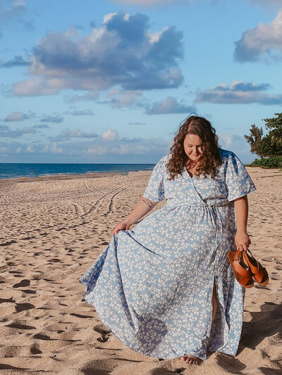 Plus size photographer poses in a blue dress holding leather sandals on the beach in Hawaii