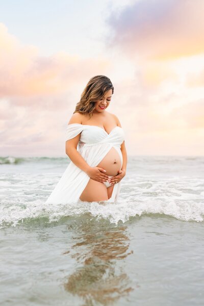 Maternity portrait of woman in white gown with exposed belly looking down and holding her baby bump and waves splashing around her.