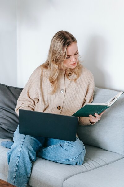 A blond woman looks down at a book while holding a laptop in her lap