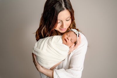 dark hair charlotte mother wearing cream holds her newborn daughter wrapped in cream on a nuratral studio background
