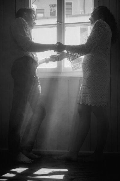 Sonnenstrahlen scheinen durch das Fenster, vor dem ein Händchen haltendes Paar steht und sich ansieht.