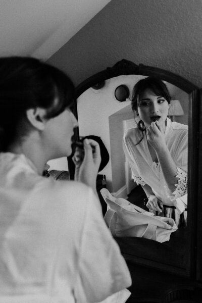 bride getting ready in front of mirror wearing a robe, she is applying lipstick