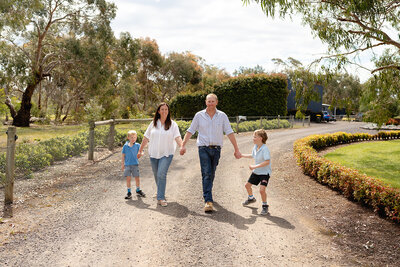 Family photography in rural Geelong showing parents walking hand in hand with their two young sons along a country driveway surrounded by trees and gardens.
