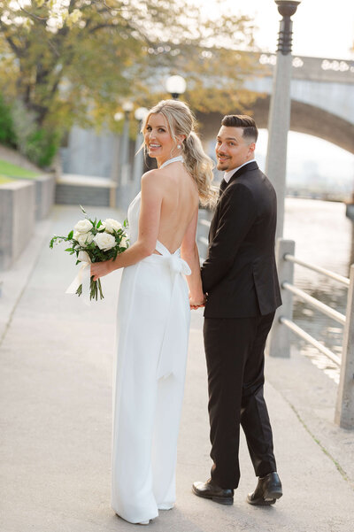 a photo of a bride in a white pantsuit and a groom in a black suit walking hand in hand and looking back smiling taken outside the NAC by Ottawa wedding photographer JEMMAN Photography