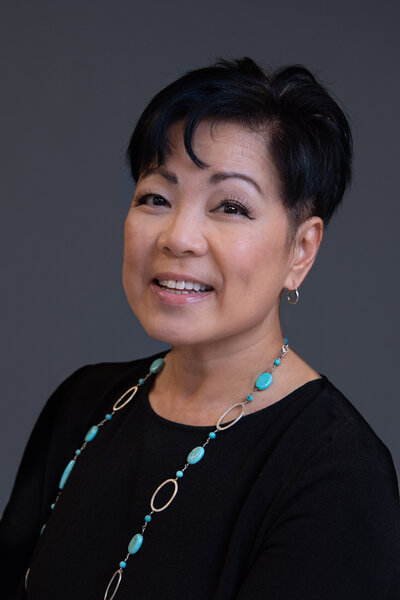 An Asian woman with a turquoise necklace and a black dress smiling and posing in front of a gray backdrop for her headshot photoshoot.